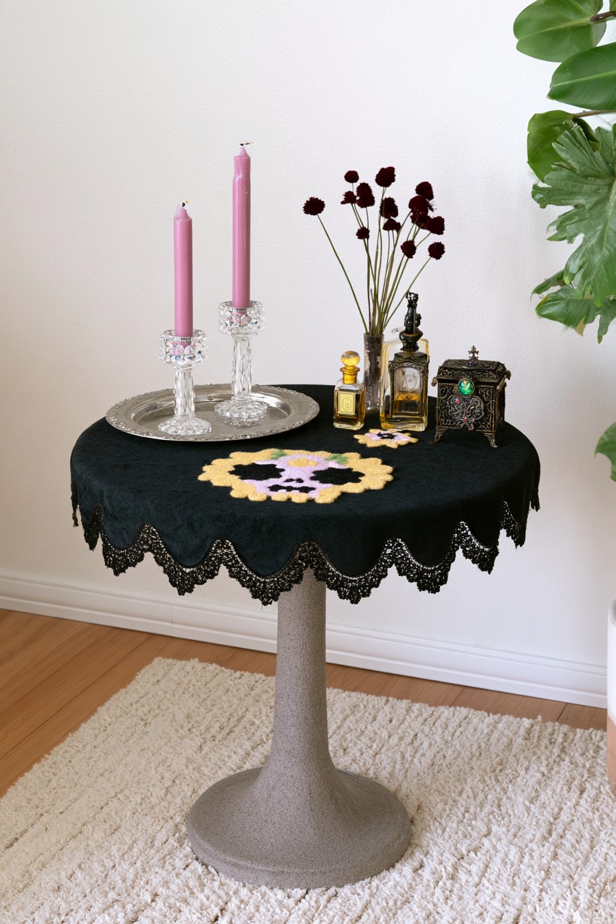 Round table with a black lace edged cloth, colorful skull motif, pink tapers, perfume bottles, and dark flowers
