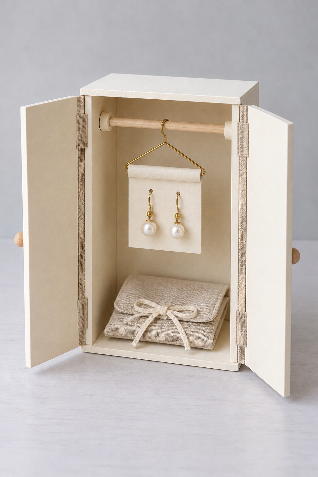 Open cream miniature wardrobe with pearl earrings on a hanging card and a linen pouch underneath