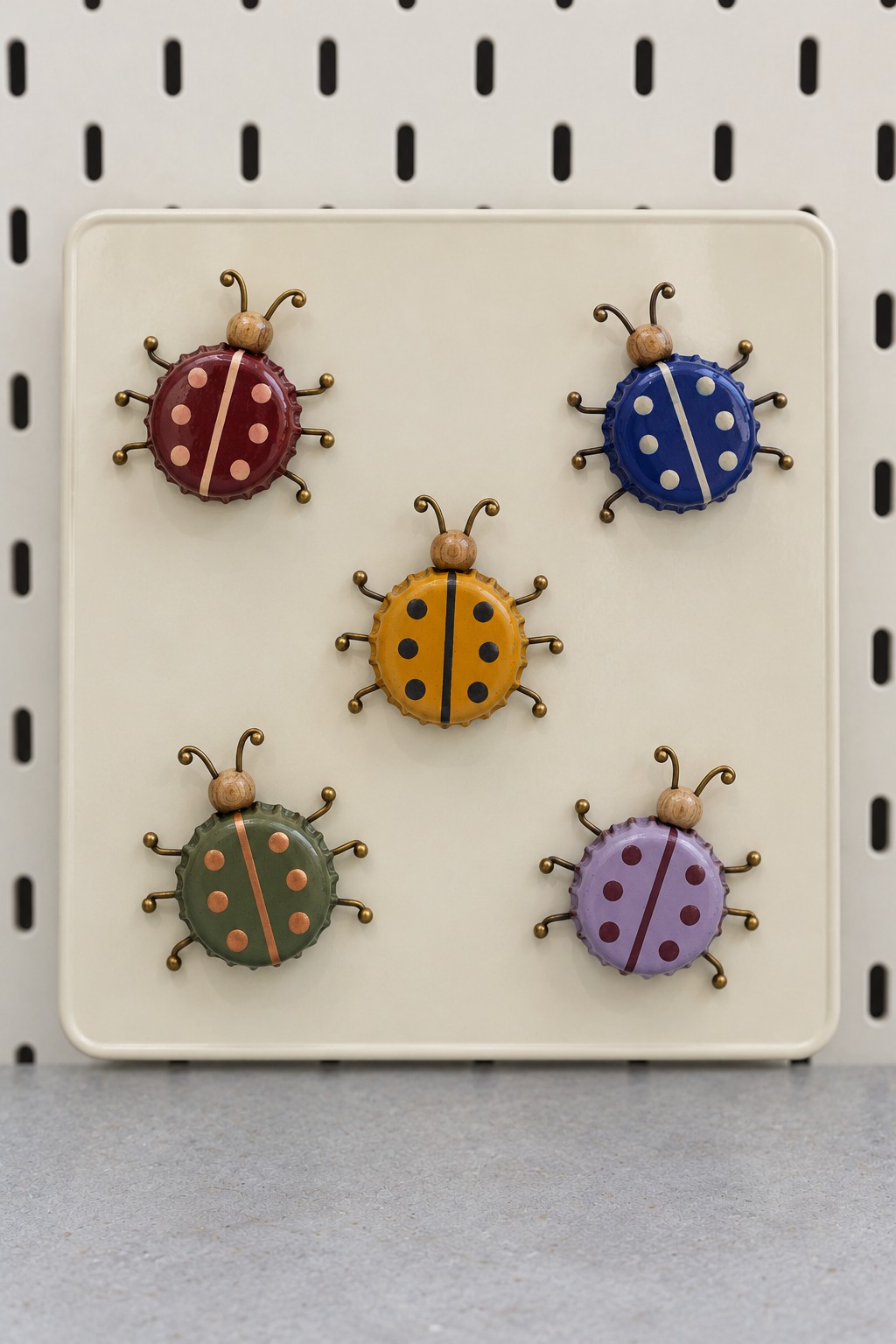 Five painted bottle cap ladybugs in burgundy, blue, yellow, green, and lilac with curled wire legs on a cream panel