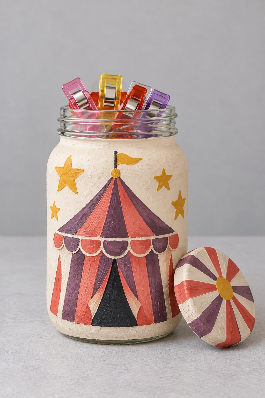 Cream jar painted with a burgundy and coral circus tent gold stars and matching striped lid holding colorful clips