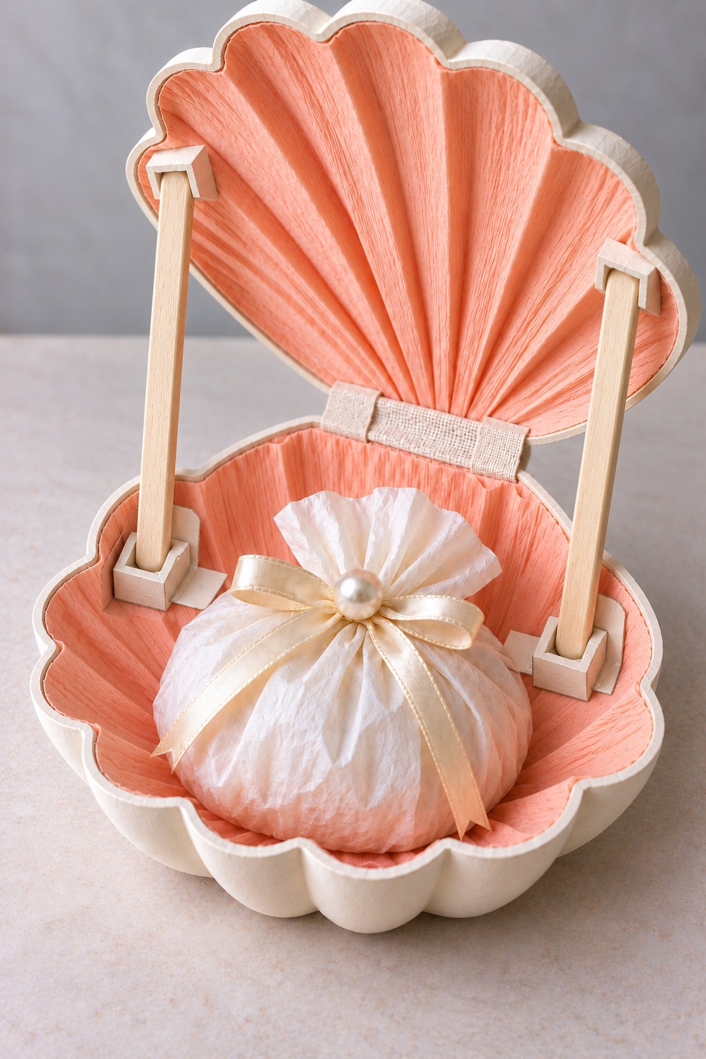 Open cream shell box with coral crepe lining, two wooden props, and a tissue parcel tied with a pearl decorated bow