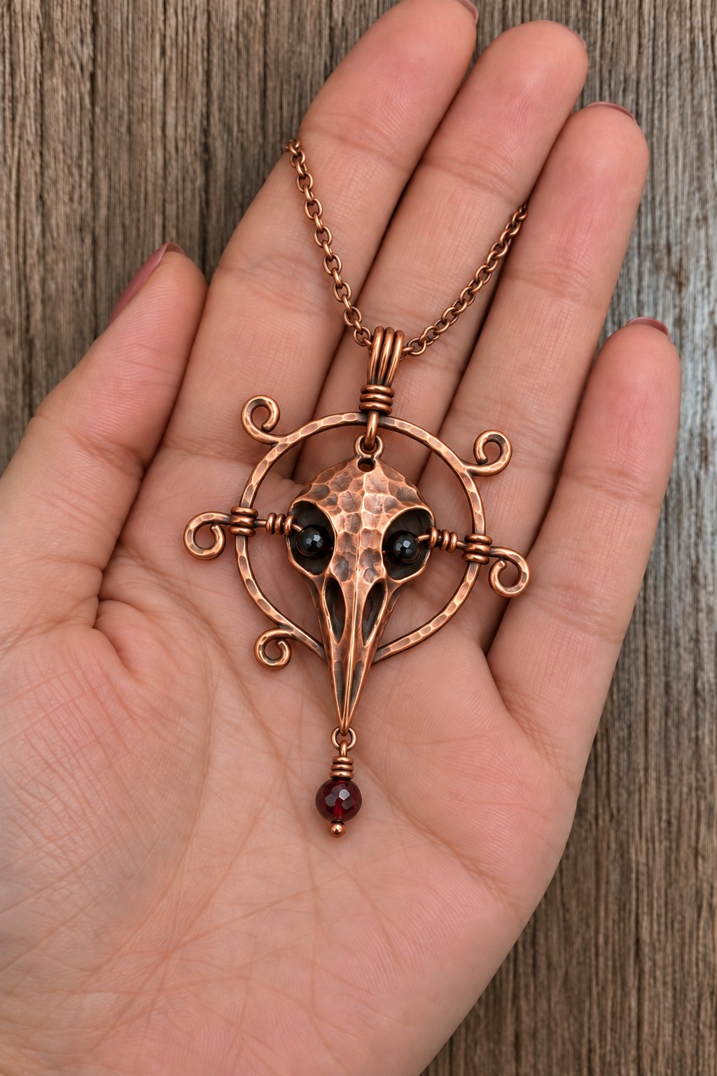 Stylized copper raven skull pendant inside a circular scrollwork frame with black eyes and a red drop
