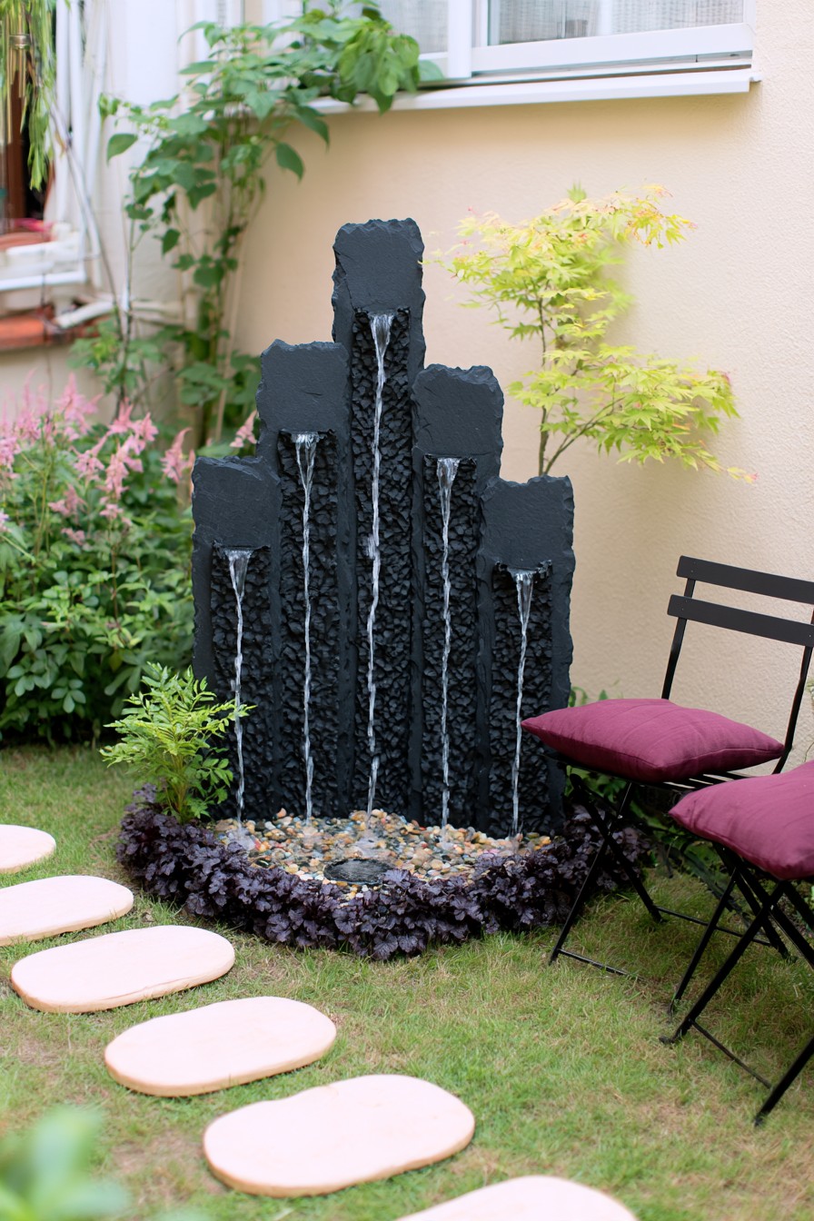 Vertical dark stone cascade with water outlets above a pebble basin beside a garden chair