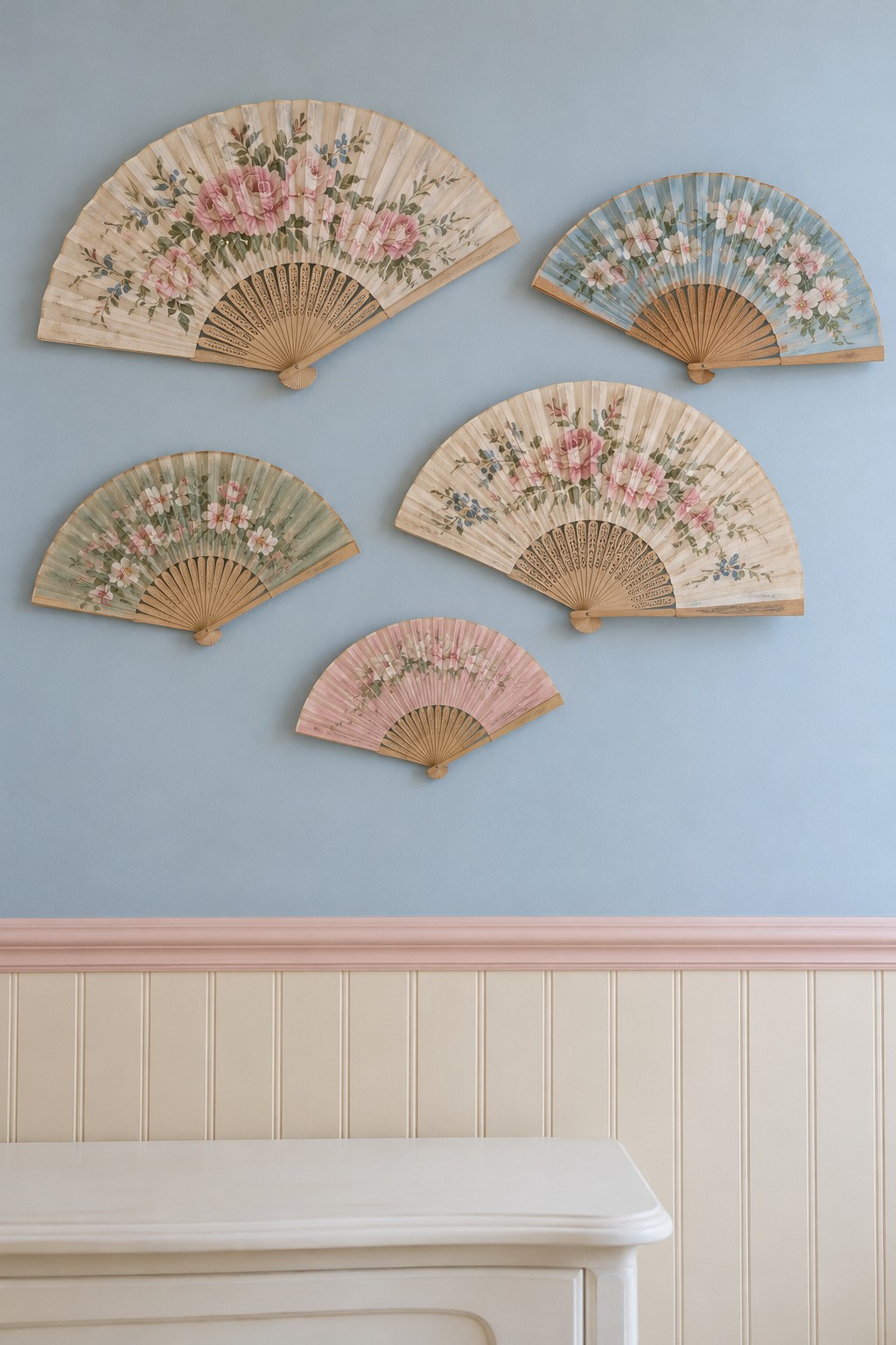Five floral folding fans arranged on a pale blue wall above cream beadboard and a pink rail