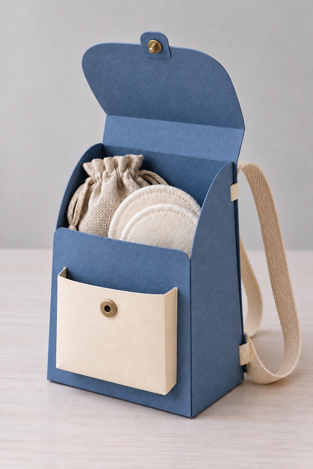 Open blue paper backpack gift bag with cream straps and pocket holding a linen pouch and reusable cotton rounds