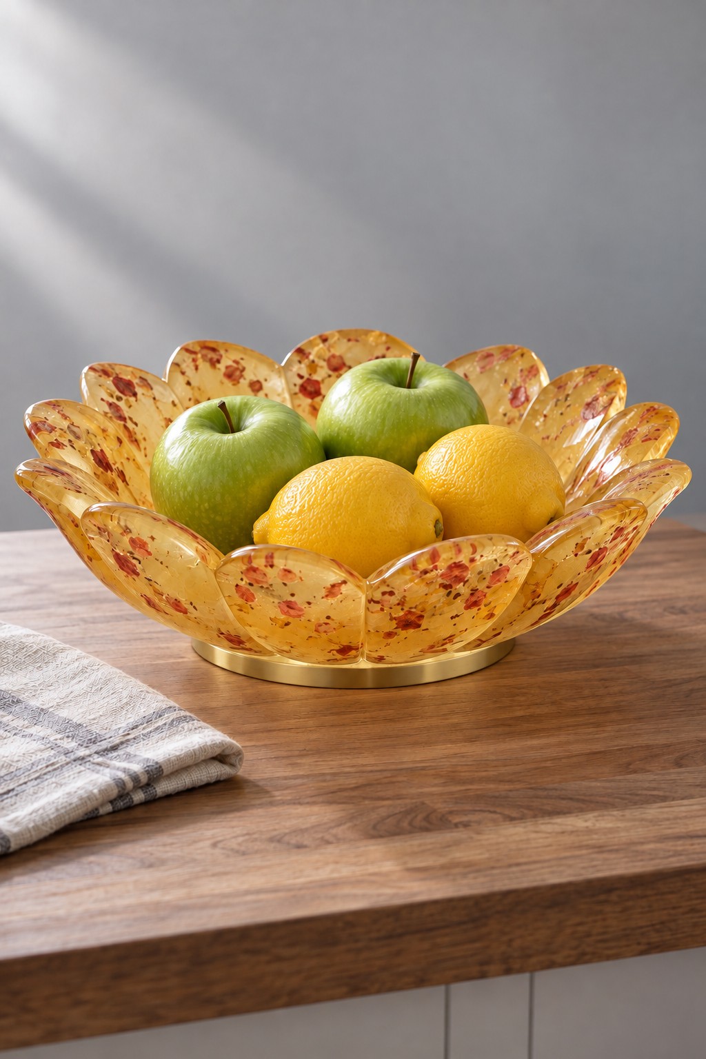 An amber and orange petal shaped resin bowl on a gold colored foot, containing green apples and yellow lemons.