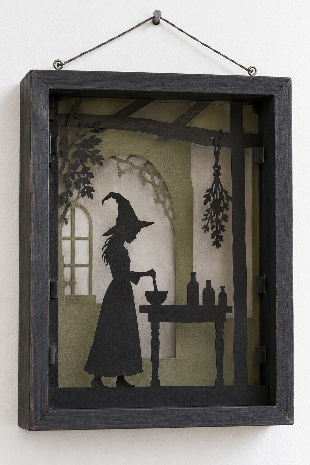 Black framed shadow box with layered paper silhouettes of a witch at a table, an arched window, shelves and hanging herbs