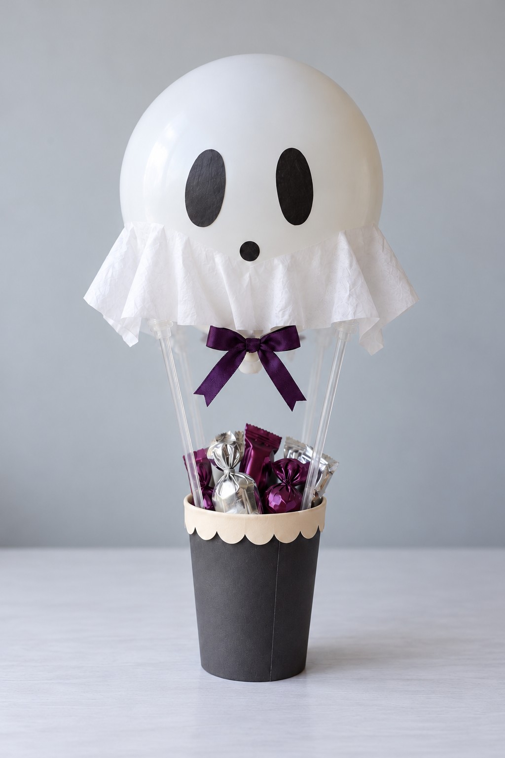 White ghost balloon with a tissue collar and purple bow above a black cup of wrapped sweets