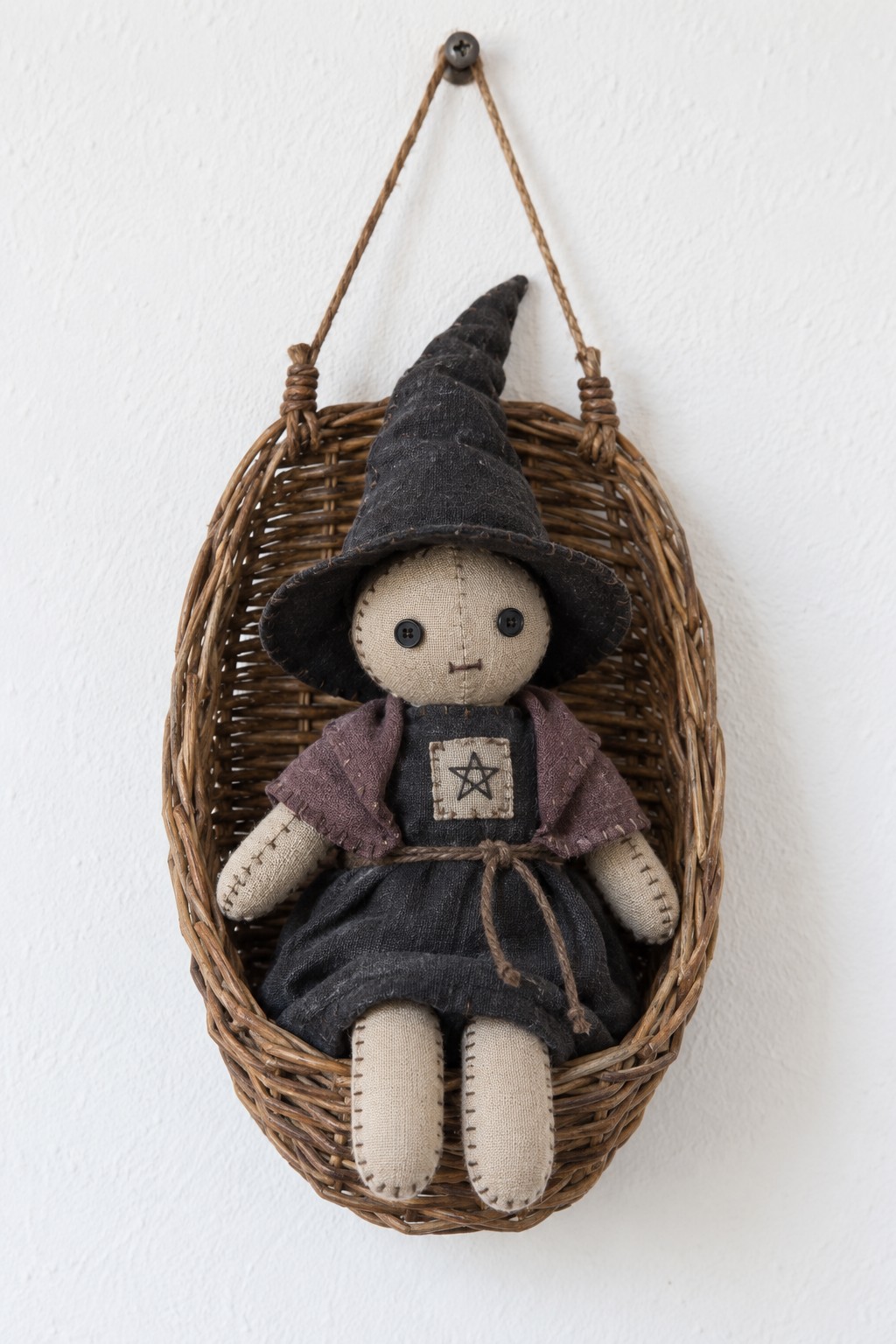 Small handmade cloth witch with a pointed black hat and plum shawl sitting inside a hanging wicker basket