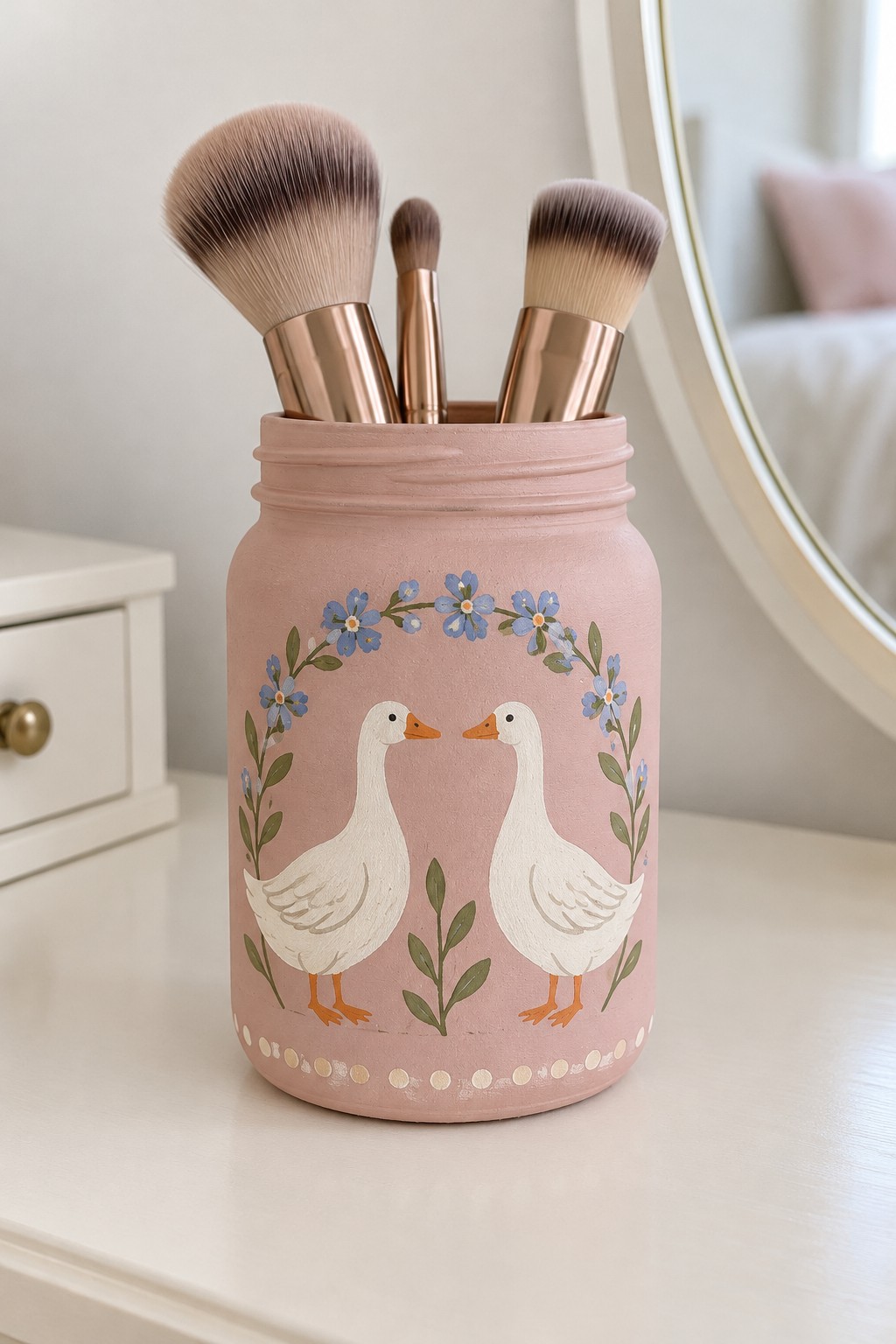 Dusty pink jar with two white geese facing each other beneath a blue floral arch holding makeup brushes
