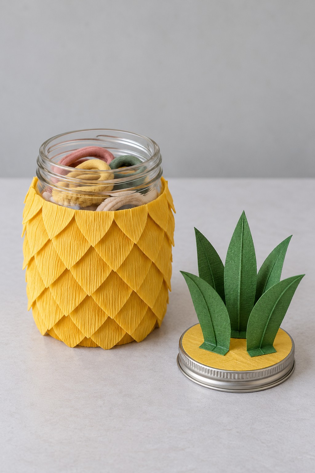 Glass hair tie jar covered with overlapping yellow paper scales beside a green leaf crown lid