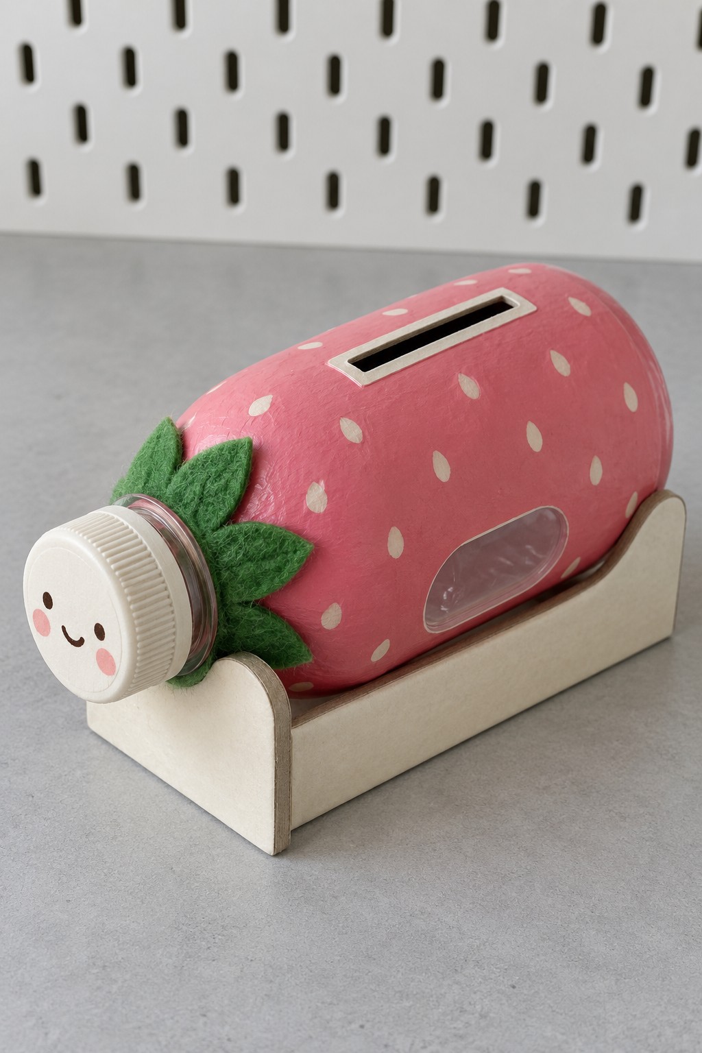 Pink strawberry bottle coin bank with green felt leaves, smiling white cap and a pale wooden cradle
