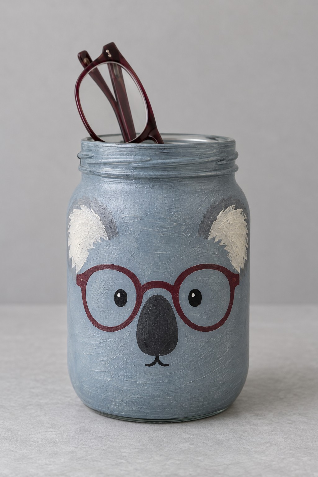 Dusty blue gray jar with a painted koala face cream ears and burgundy spectacles holding eyeglasses