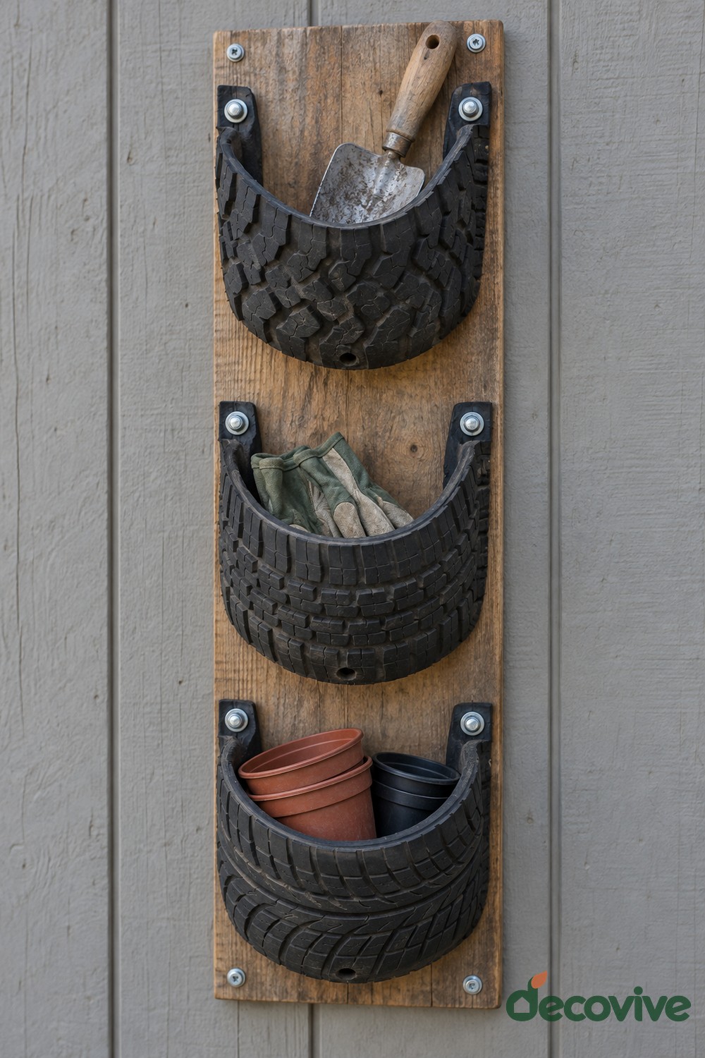 Three curved black tire pockets bolted to a vertical wooden board holding a trowel, gardening gloves, and small empty pots