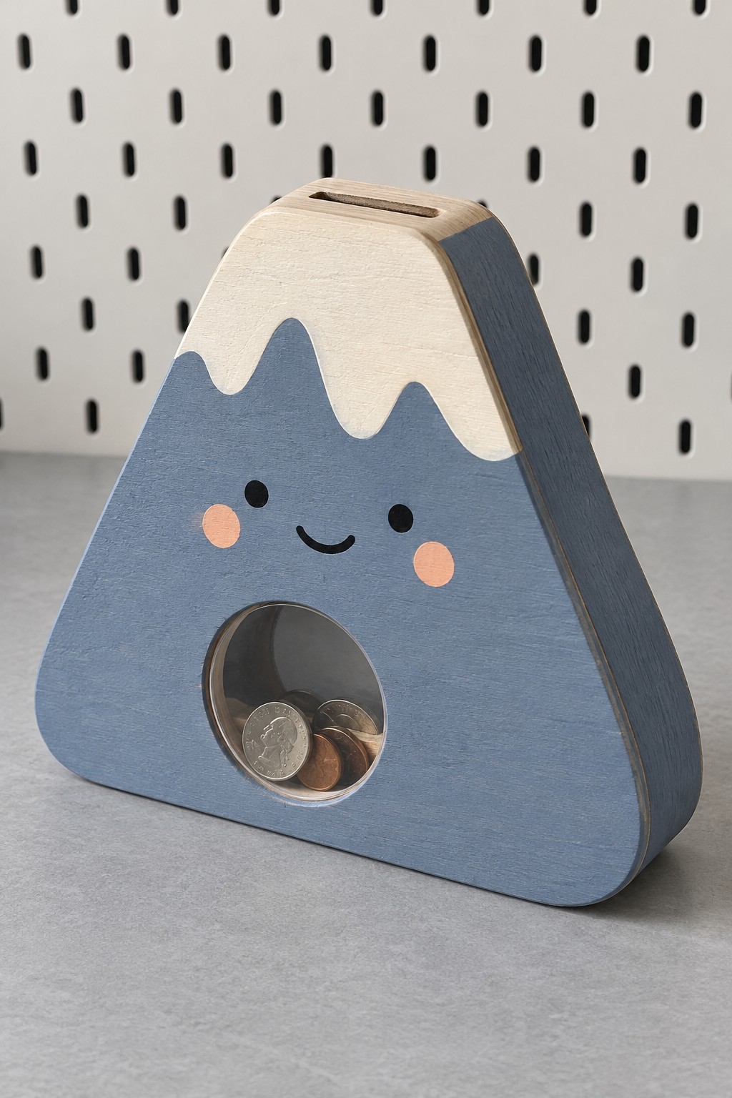 Blue mountain shaped coin bank with cream snowcap, smiling face and a circular clear window