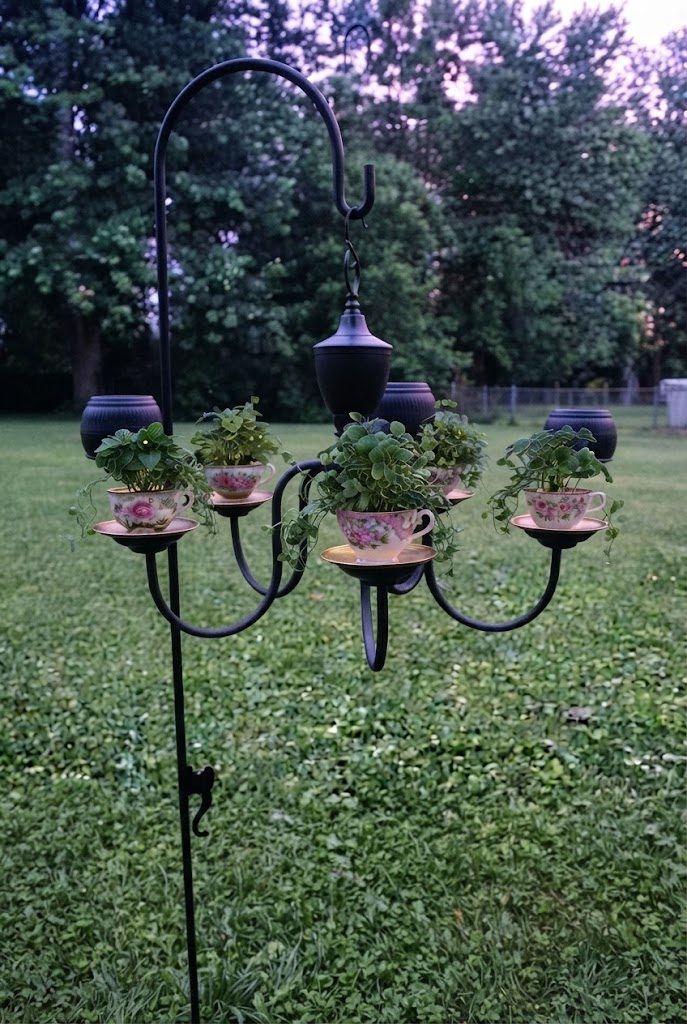 Black chandelier with floral teacups planted with green trailing plants on a shepherd's hook in a lawn