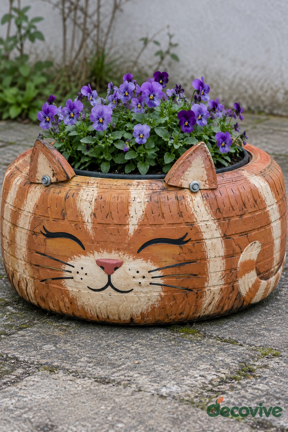 Orange and cream tire cat planter with bolted wooden ears, closed eyes, a pink nose, whiskers, and purple violas
