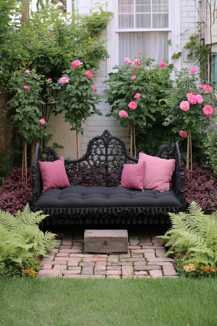 Ornate black iron garden seat with pink cushions surrounded by roses and ferns