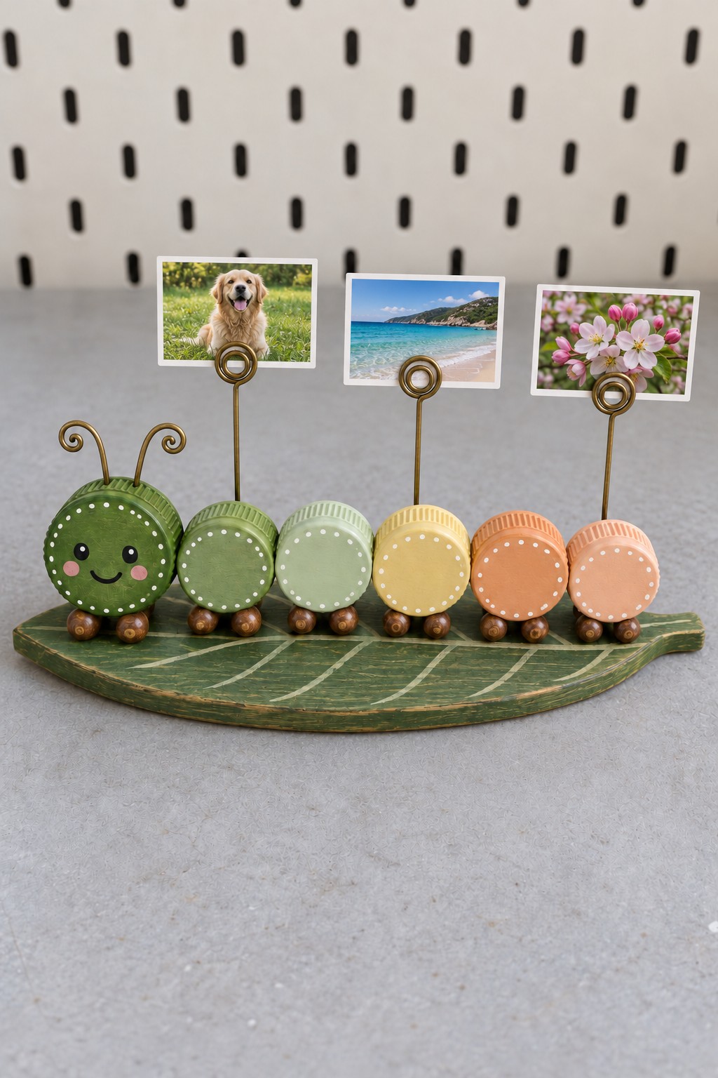 Green to peach bottle cap caterpillar with wooden bead feet and three spiral wire photo holders on a wooden leaf