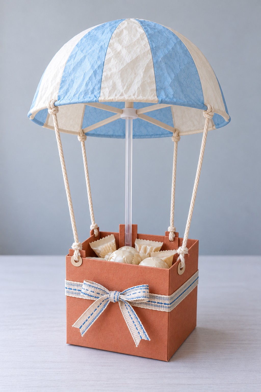 Blue and cream paper parachute on a clear central support above a terracotta sweet box with four cotton cords
