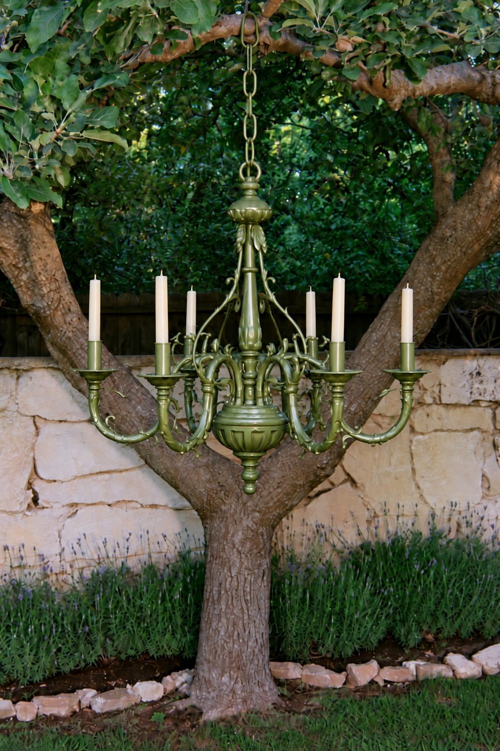 Olive green painted chandelier with white taper candles hanging from a tree branch in a garden