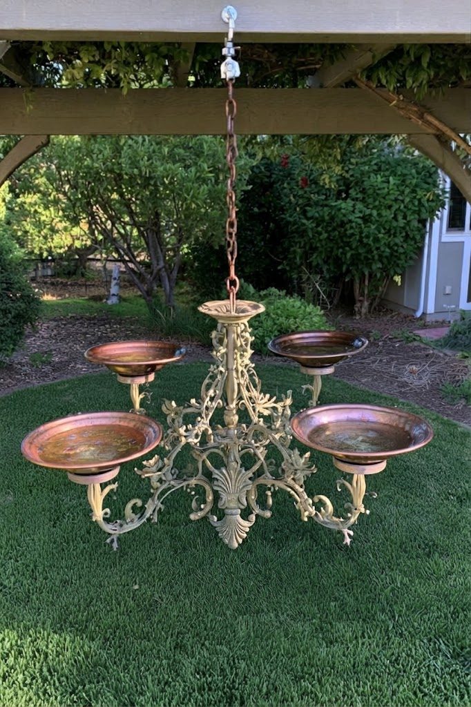 Gold painted chandelier with copper water bowls hanging from a pergola as a bird bath