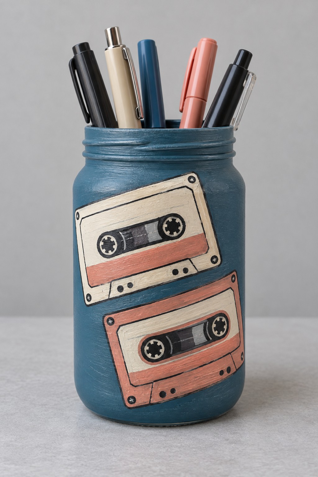 Dusty blue jar with two painted cream and coral cassette tapes holding pens