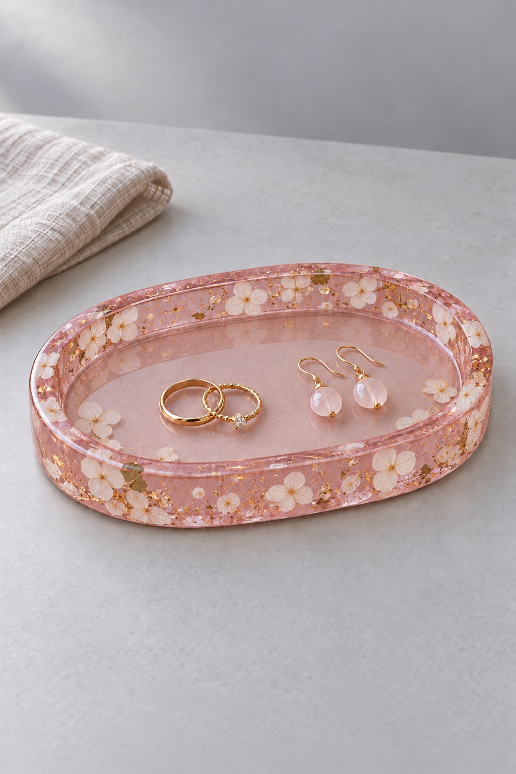 An oval blush pink resin jewelry tray with white flowers and gold flecks, holding gold rings and pale pink earrings.