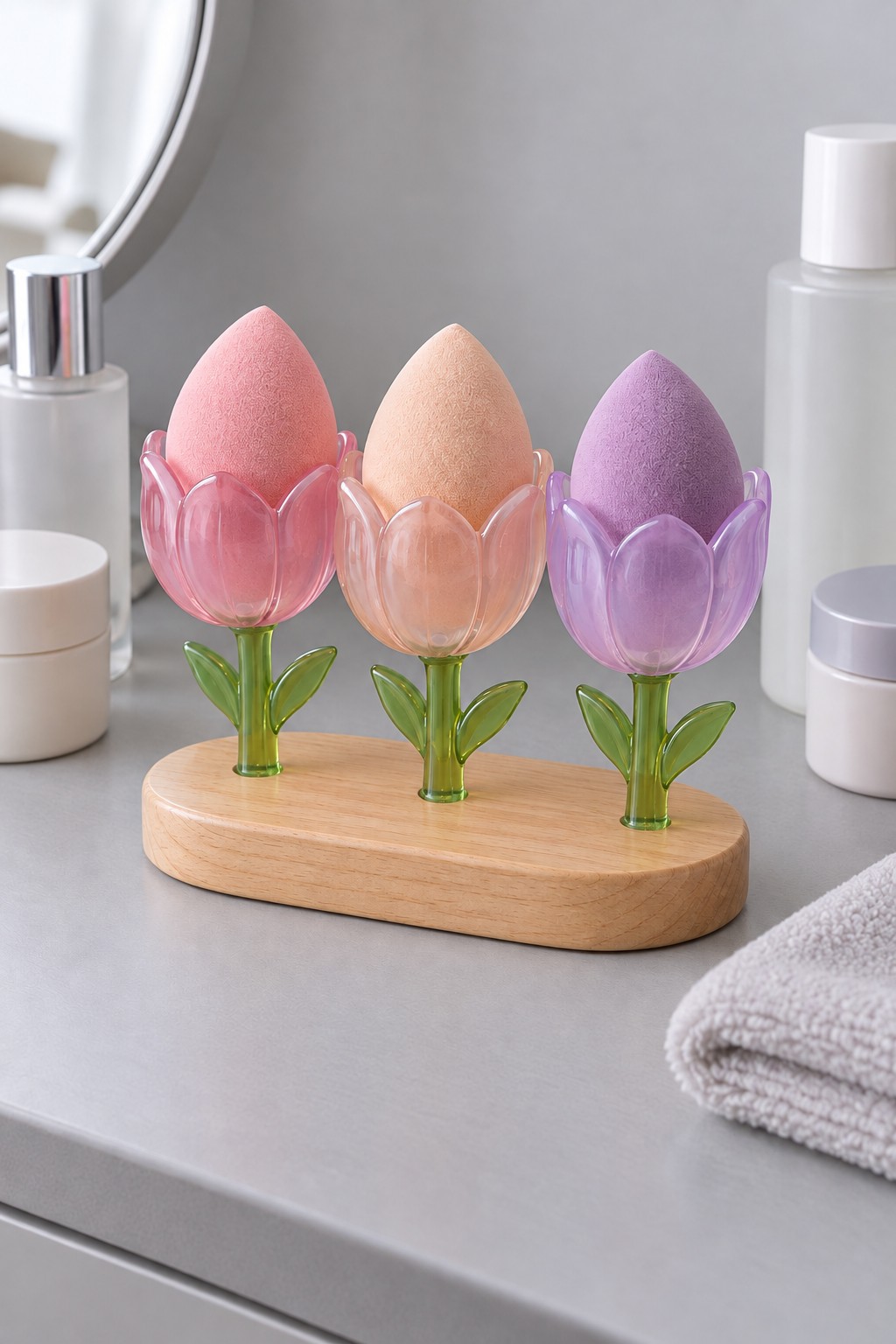 Three pink, peach and lilac tulip shaped resin cups with green stems on an oval wooden base, holding makeup sponges.