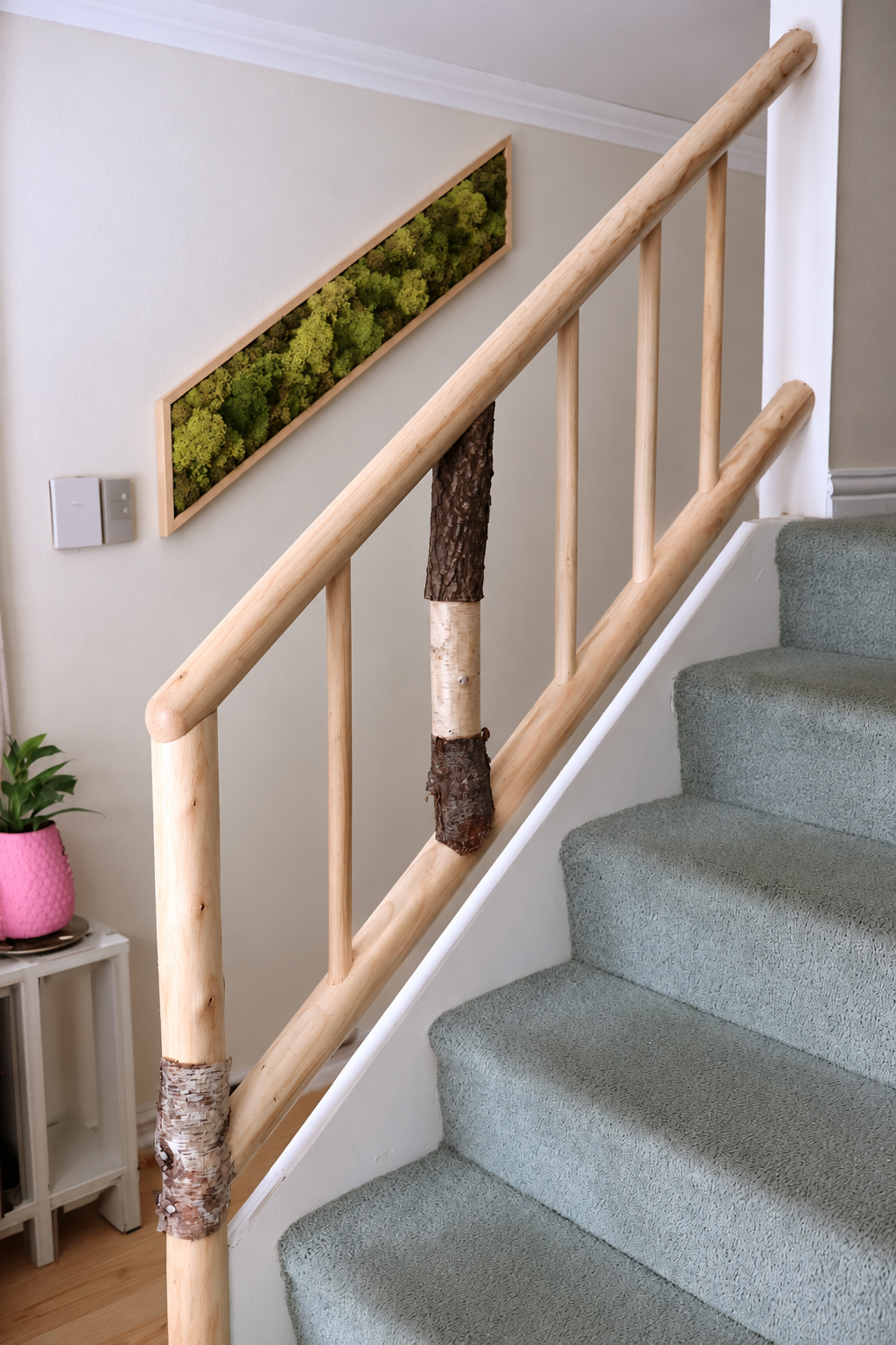 Bare wooden stair handrail with birch accented uprights beside a separate framed moss wall panel