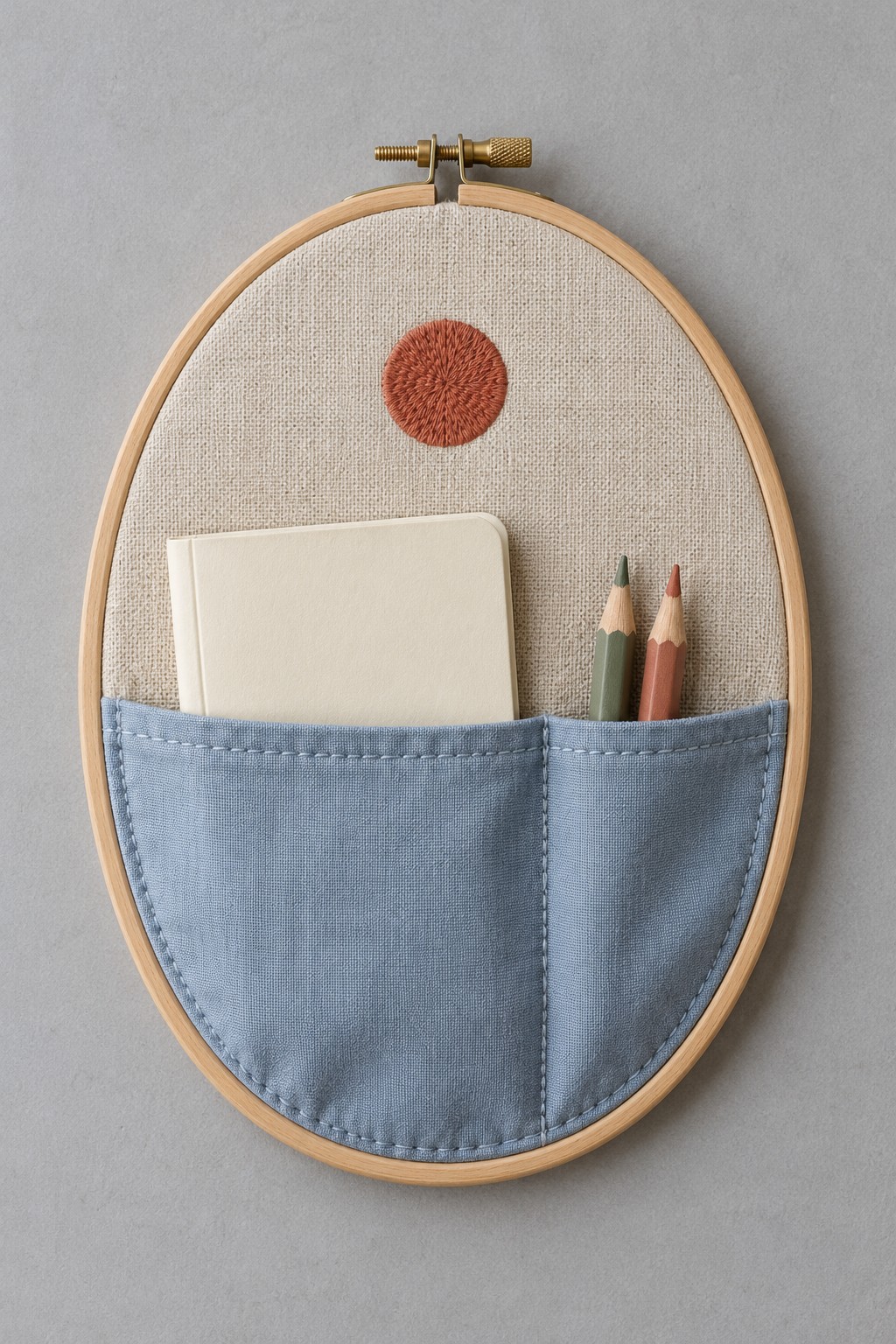 Oval wooden embroidery hoop with linen backing, a blue divided pocket, a notebook, and two pencils