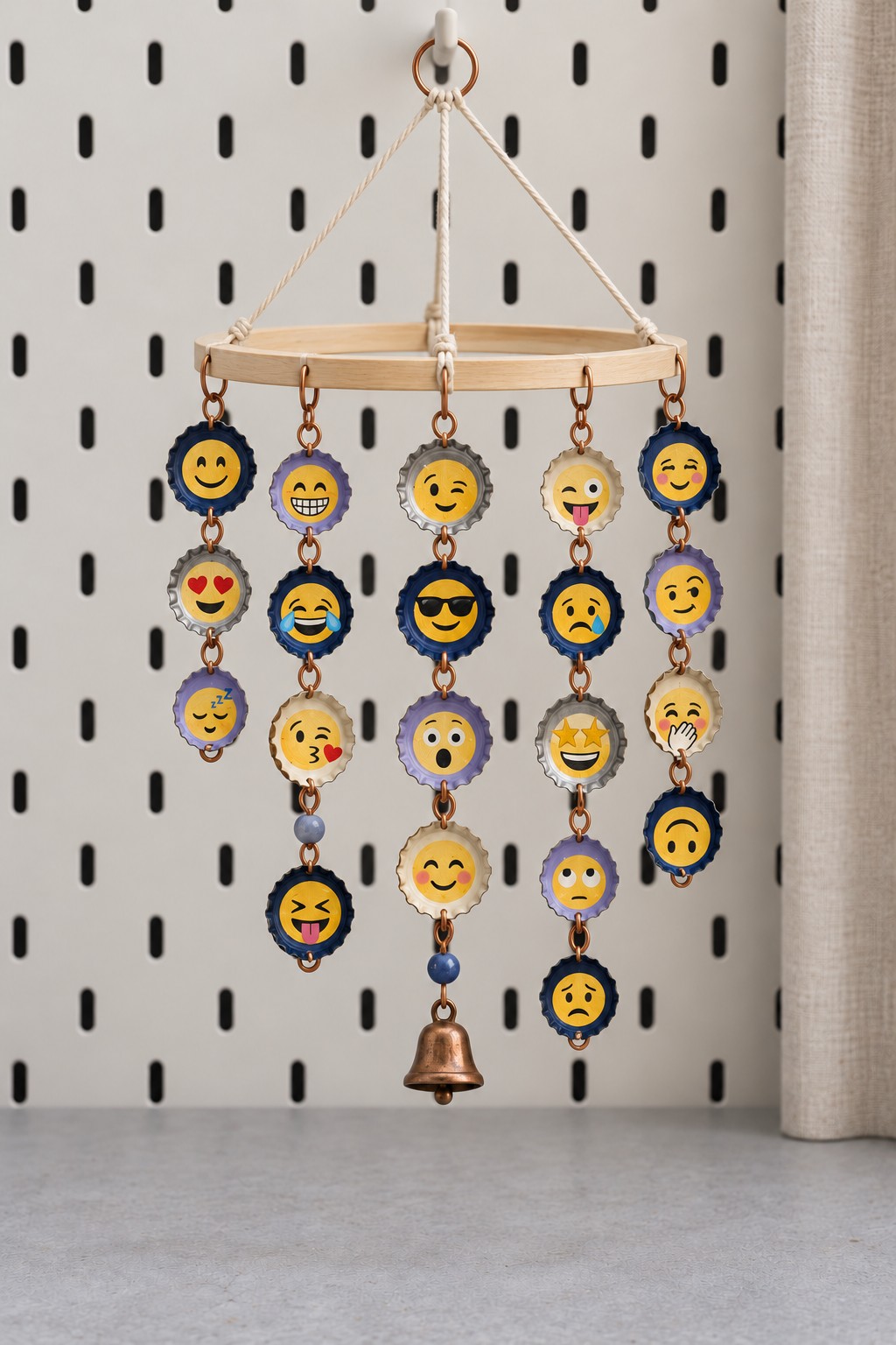 Five hanging strands of painted emoji bottle caps linked with copper colored rings beneath a wooden hoop with a small bell