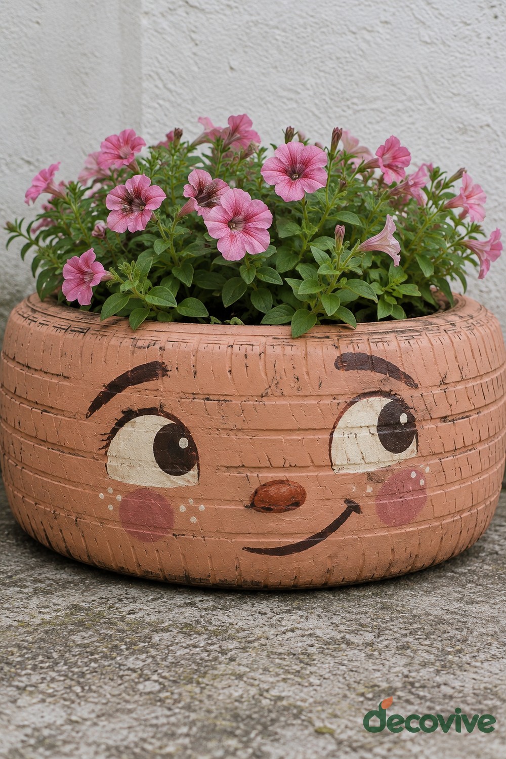 Peach tire planter painted with wide eyes, raised eyebrows, rosy cheeks, and a smile beneath pink petunias