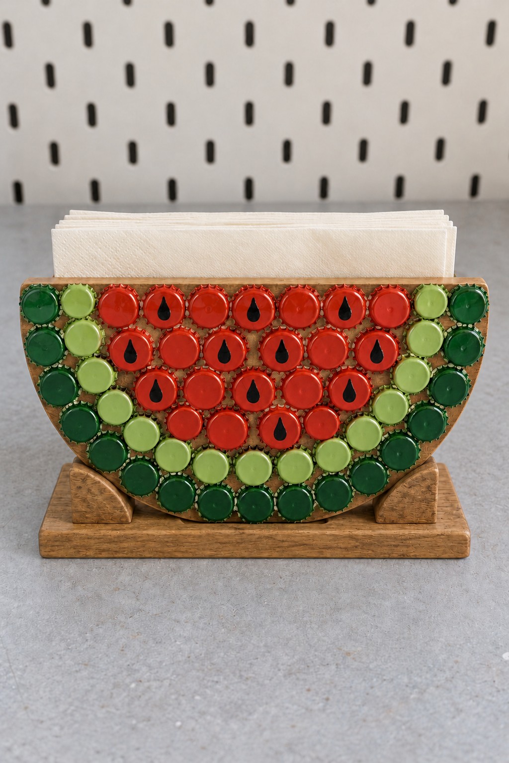 Watermelon slice napkin holder made with red and green metal bottle caps and black seed details on a wooden base