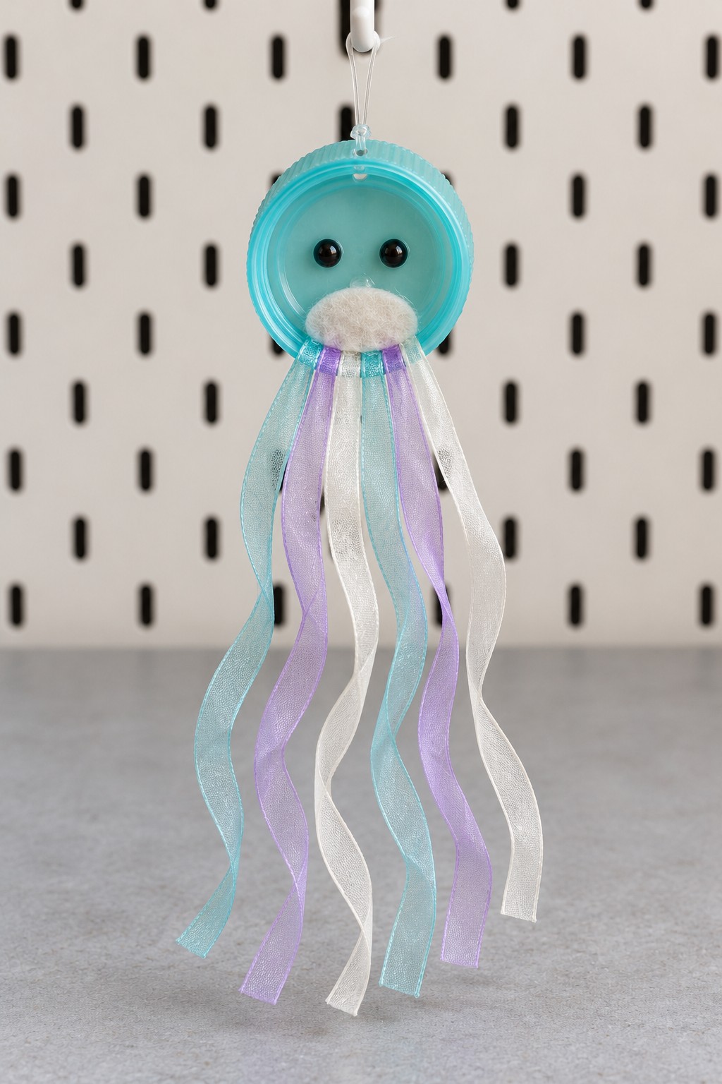 Turquoise plastic cap jellyfish ornament with black eyes, a pale mouth detail, and long mint, lilac, and white ribbon tentacles