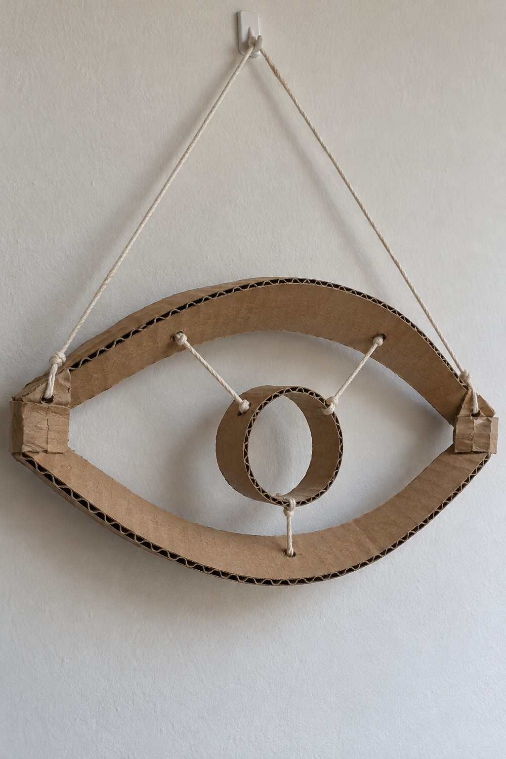 Eye shaped corrugated cardboard sculpture with a central oval ring held by three cords