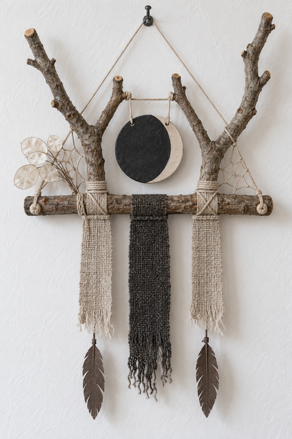 Two forked branches tied to a crossbar with a black and cream moon disc, woven fabric strips and two feather pendants
