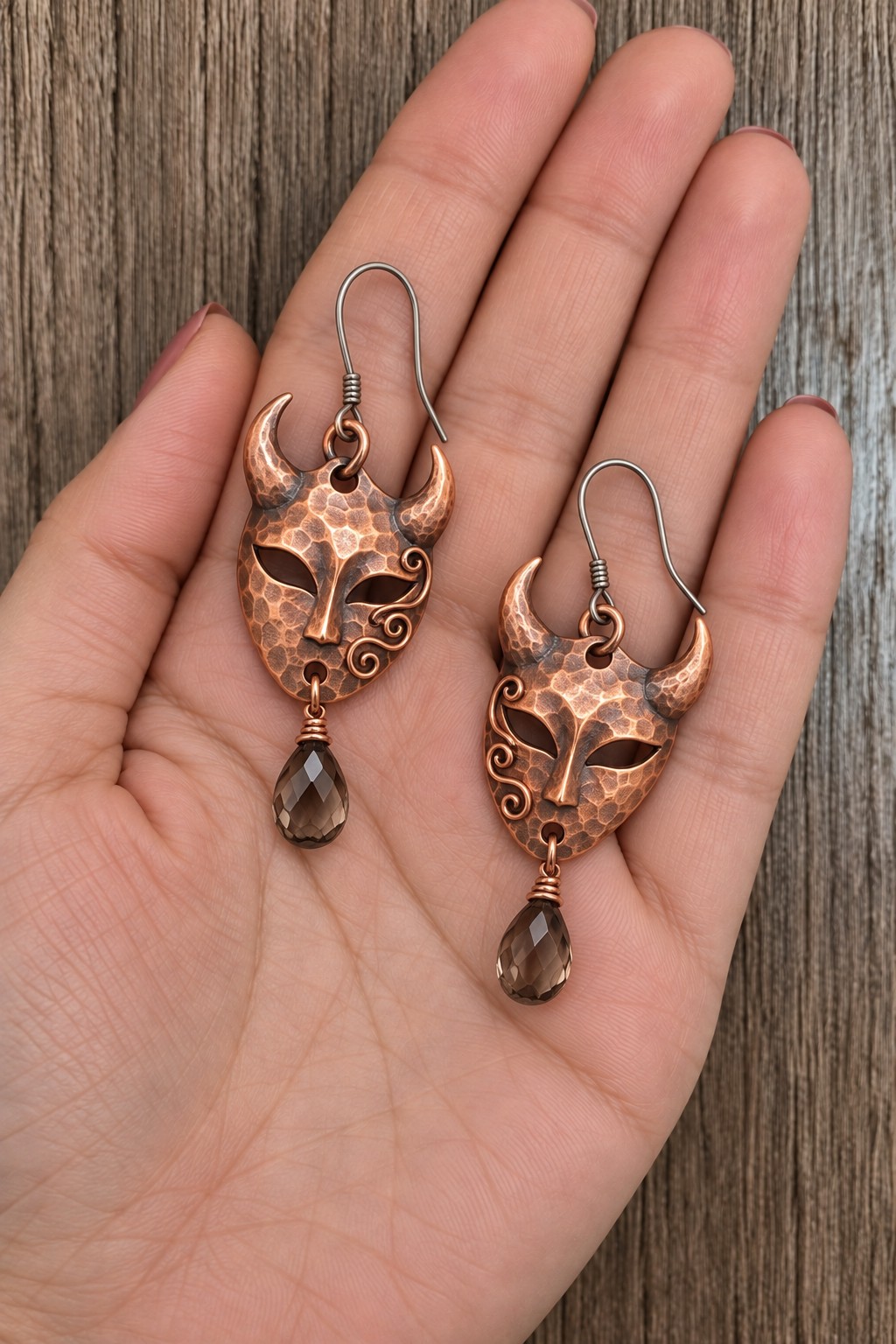 Pair of horned copper mask earrings with open eyes, spiral details, and smoky teardrop beads