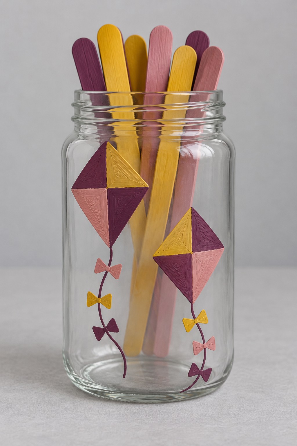 Clear jar painted with two mustard burgundy and dusty pink kites with bow tails holding matching colored craft sticks
