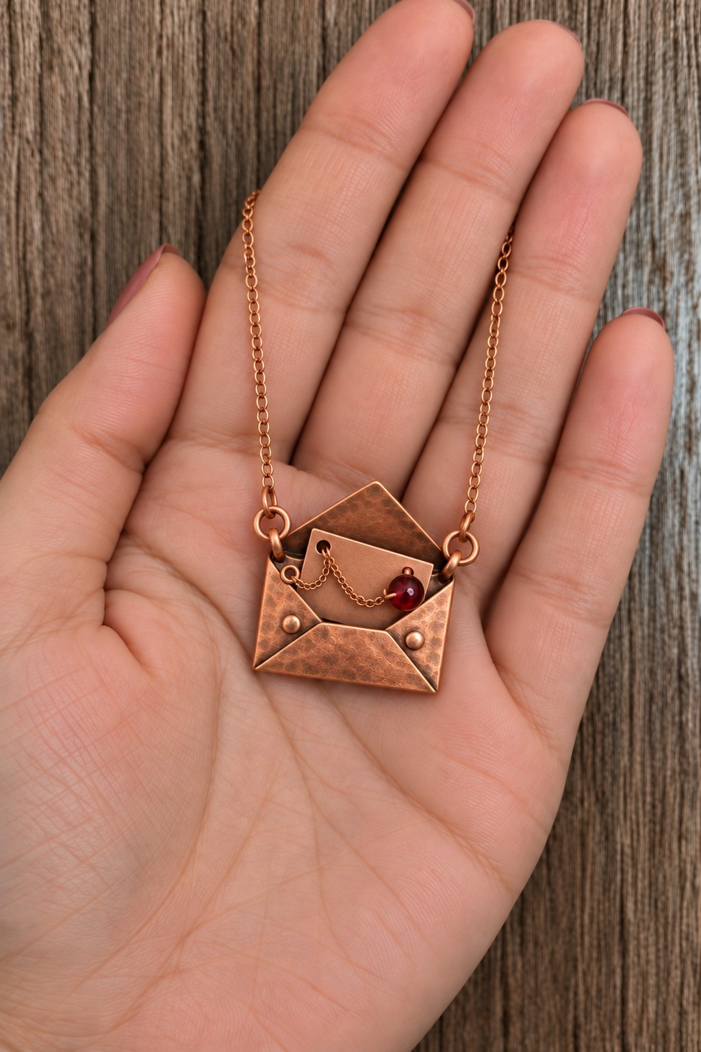 Small copper envelope pendant containing a metal letter and a red bead on a short chain