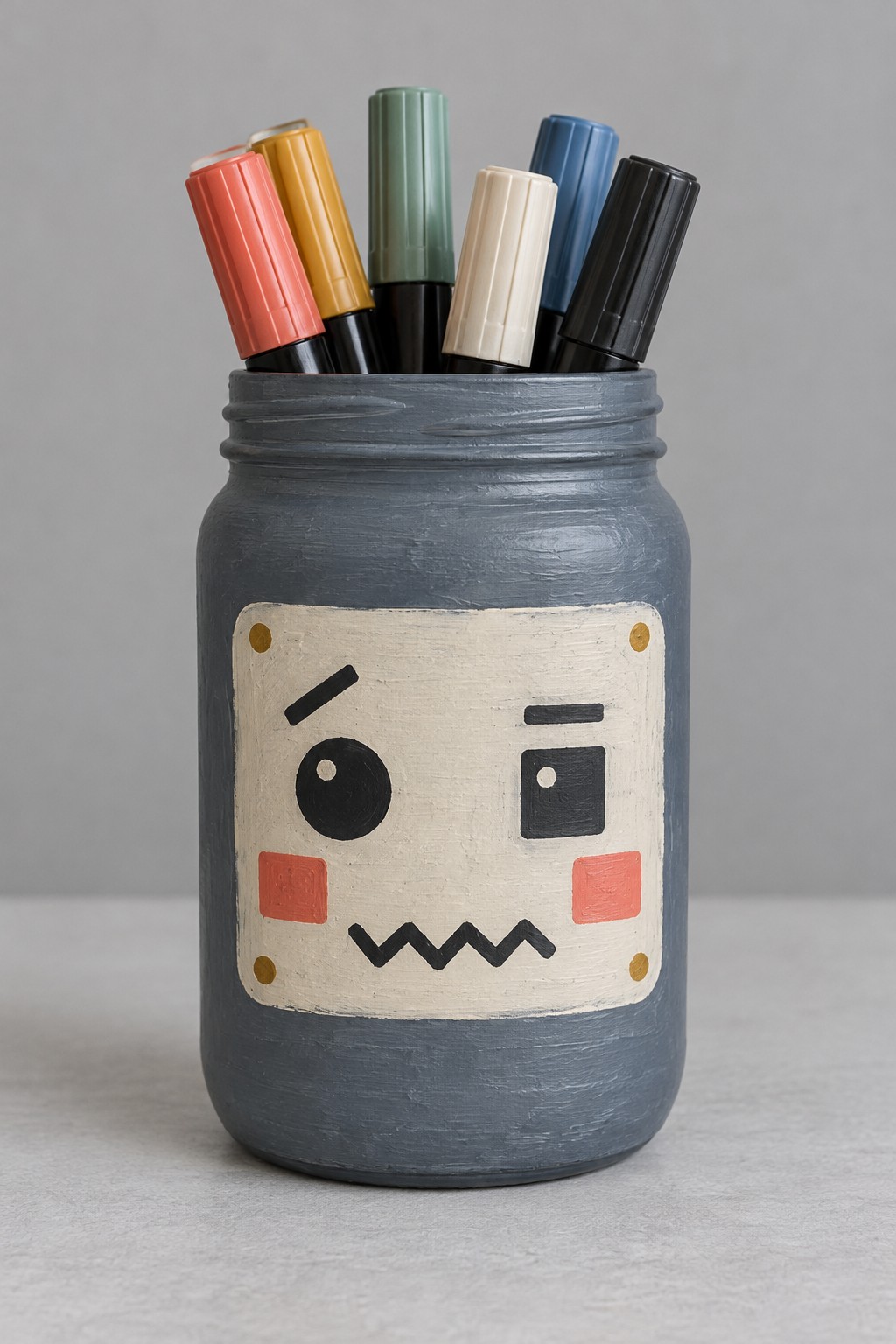 Charcoal gray jar with a cream robot face panel and mismatched geometric eyes holding markers