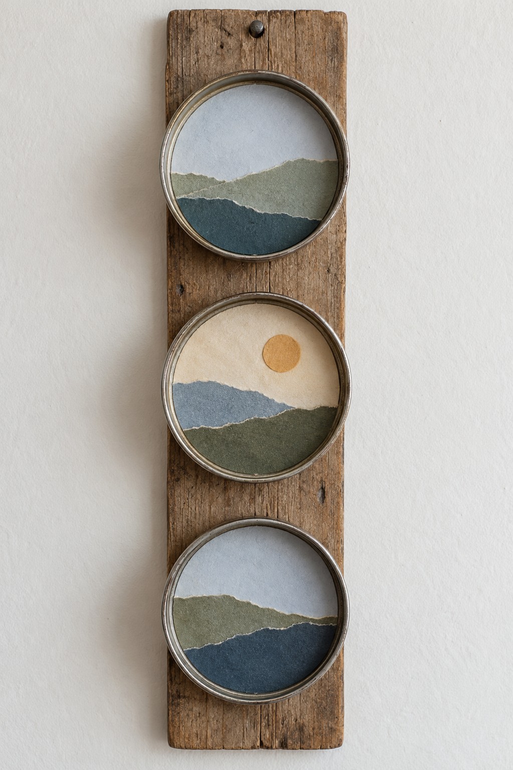 Three circular torn paper landscapes in metal lids arranged vertically on a weathered wooden board