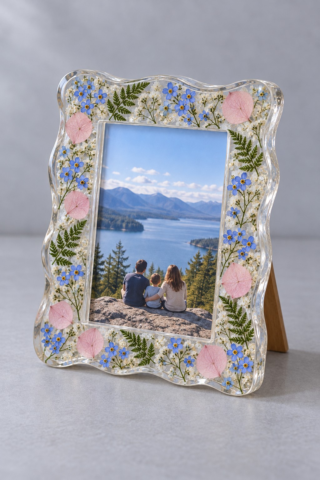 A clear wavy resin photo frame containing ferns, small blue flowers and pink petals around a landscape photograph.