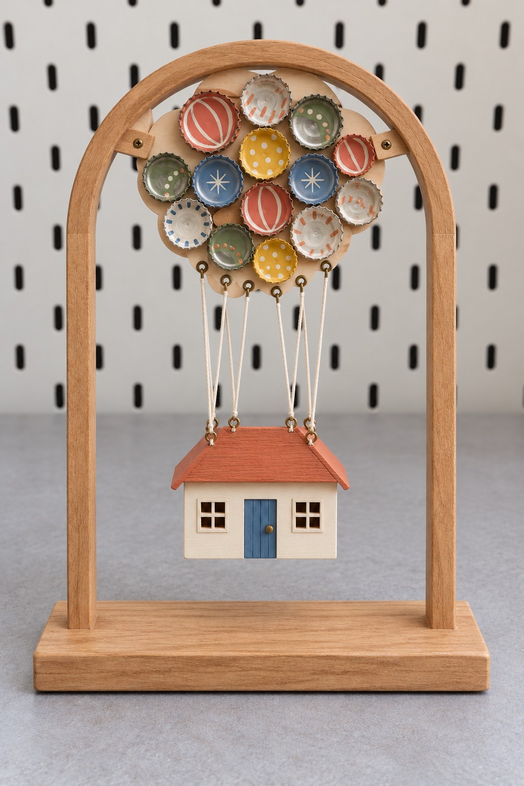 Miniature cream house suspended by cords below a cluster of painted metal bottle caps inside a wooden arch