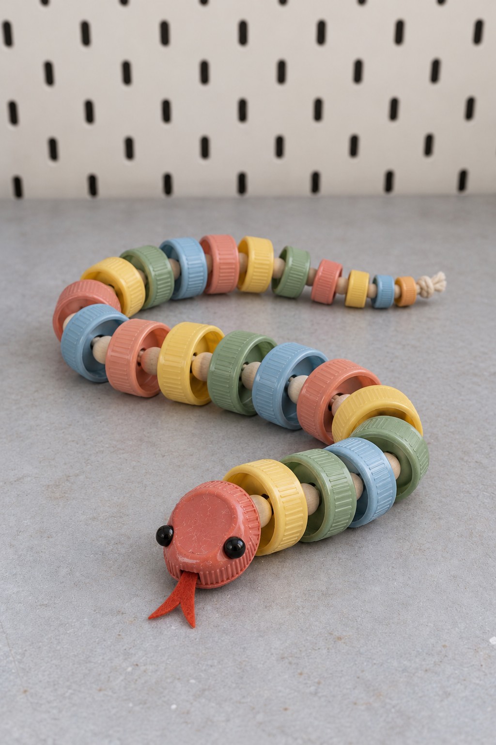Long pastel plastic bottle cap snake threaded on cream cord with a coral head, black eyes, and red felt tongue