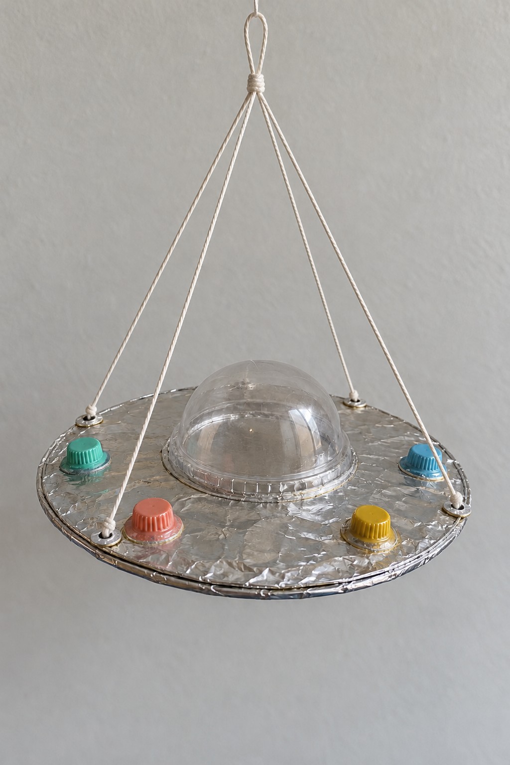 Silver foil covered flying saucer ornament with a transparent dome colored bottle caps and three hanging cords