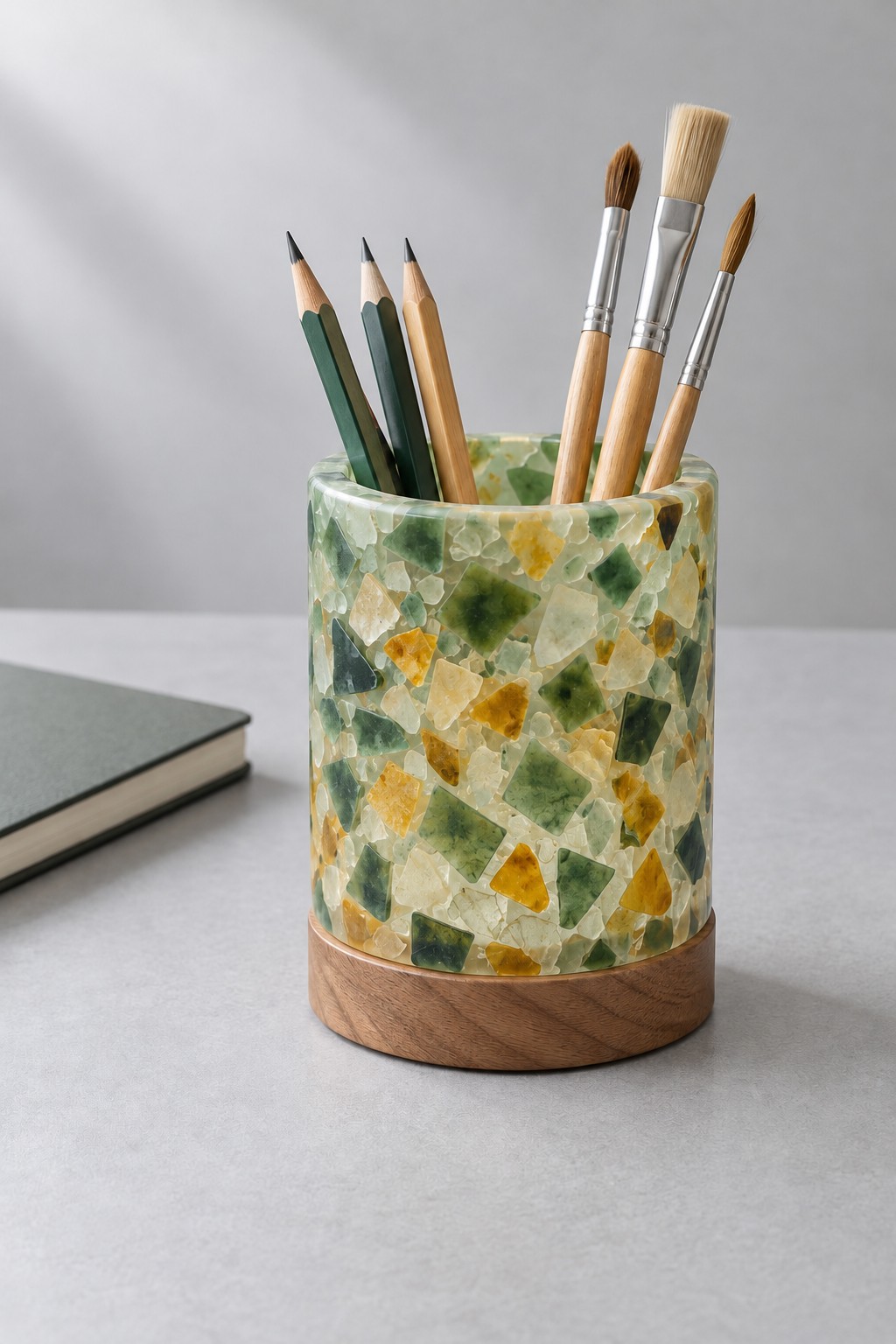 A cylindrical green, yellow and clear mosaic effect resin cup on a wooden base, holding pencils and paintbrushes.